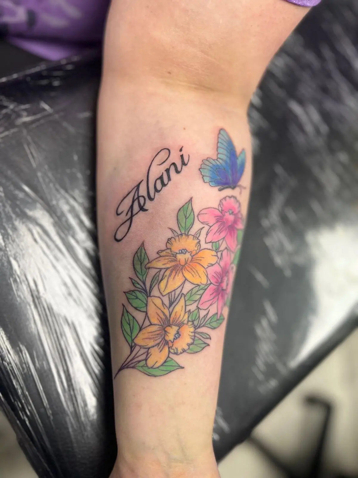 Forearm memorial tattoo with the script name Alani, a blue butterfly and yellow daffodils with pink flowers, by Carly
