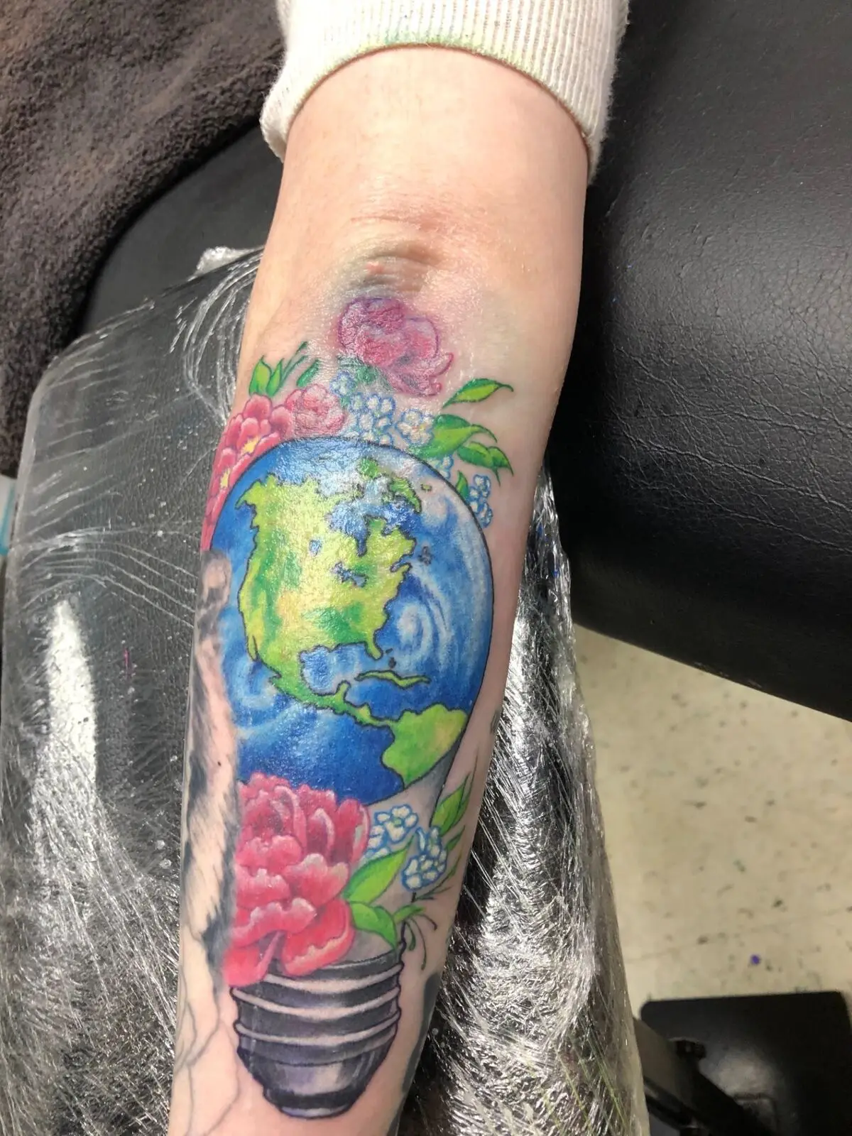 Color earth-in-a-lightbulb tattoo with peonies and forget-me-nots on forearm