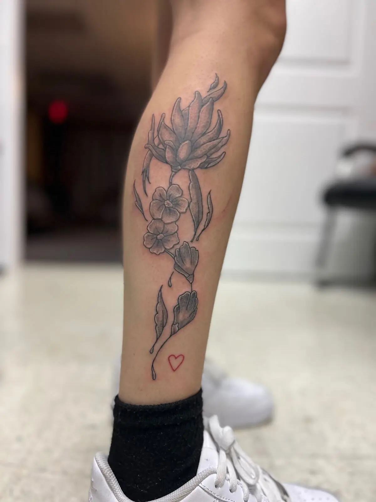 Black and grey floral calf tattoo of a large blooming flower and cascading blossoms ending in a small red outlined heart, by Carly