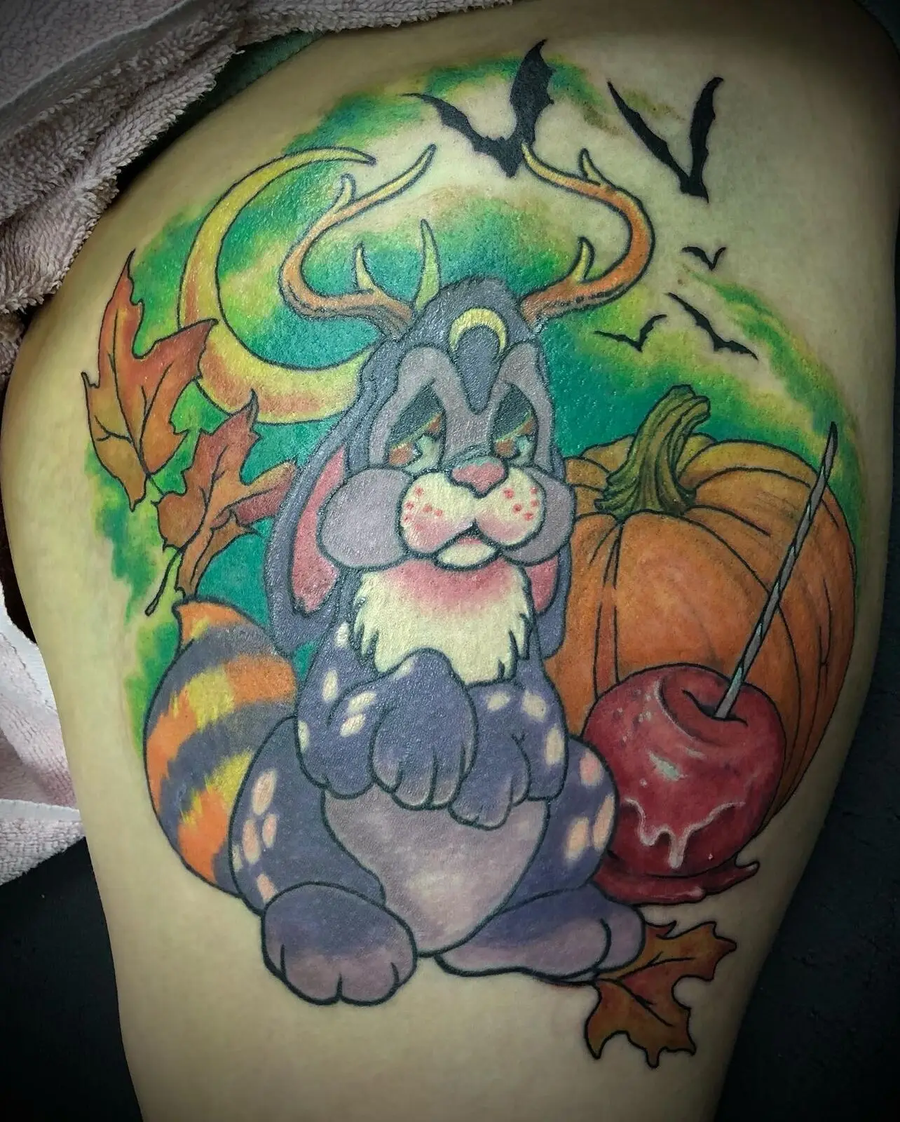 Color thigh tattoo of a cartoon antlered bunny with pumpkin, candy apple, autumn leaves and bats