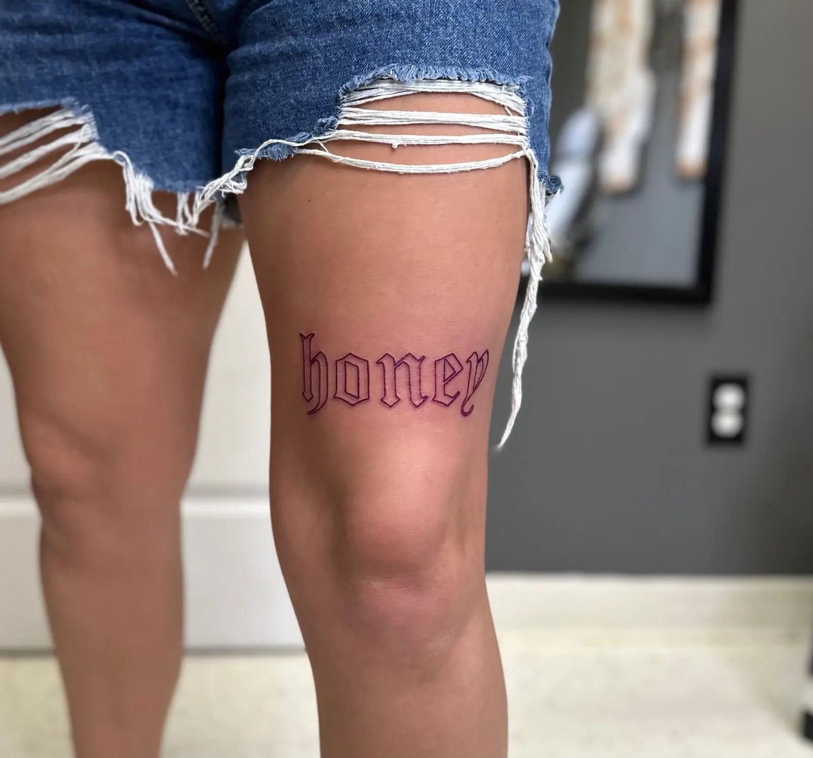 Outline of the word honey in bold gothic Old English lettering above the knee on the thigh, by Carly