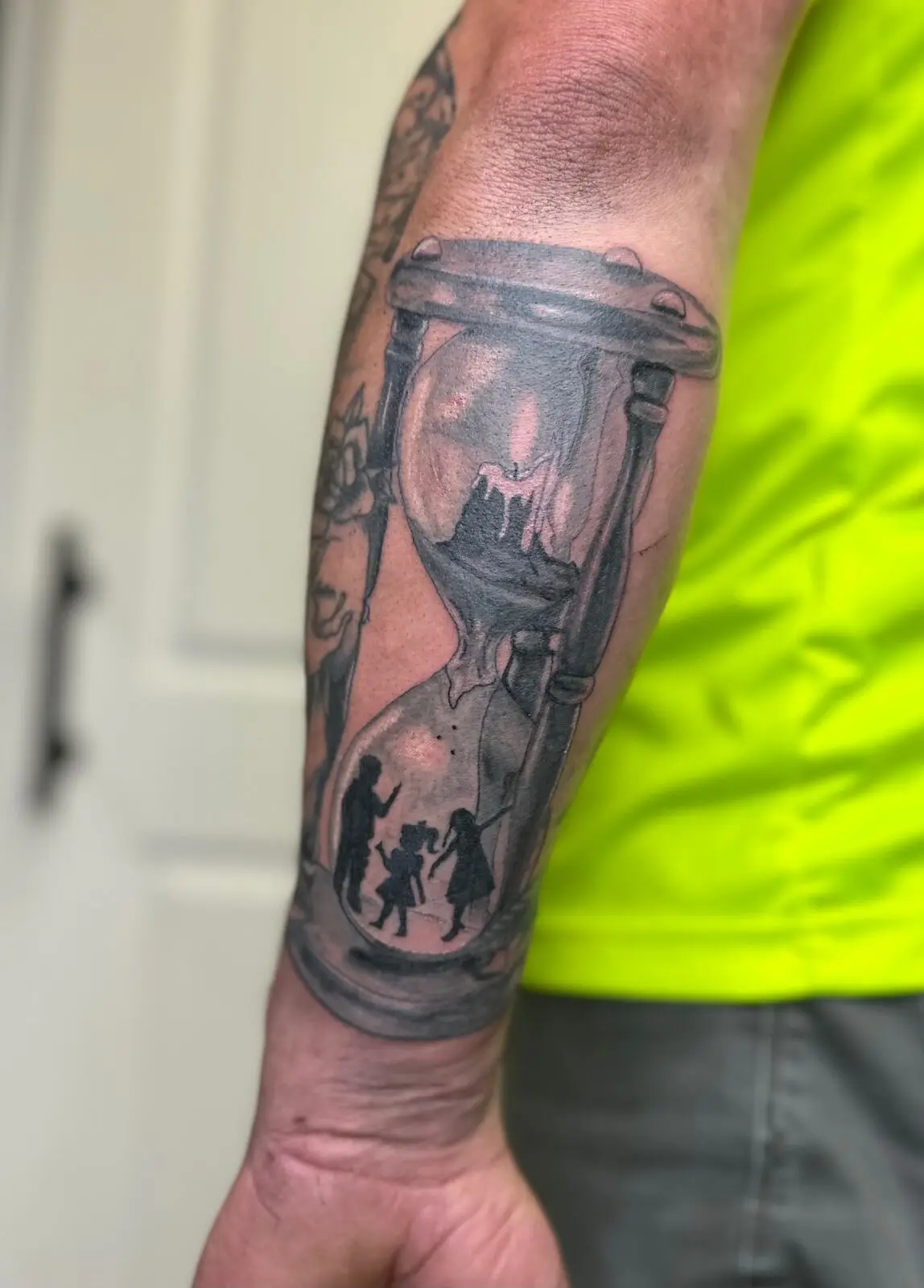 Black and grey realism forearm tattoo of an hourglass with crumbling mountain on top and family silhouettes in the bottom chamber, by Carly