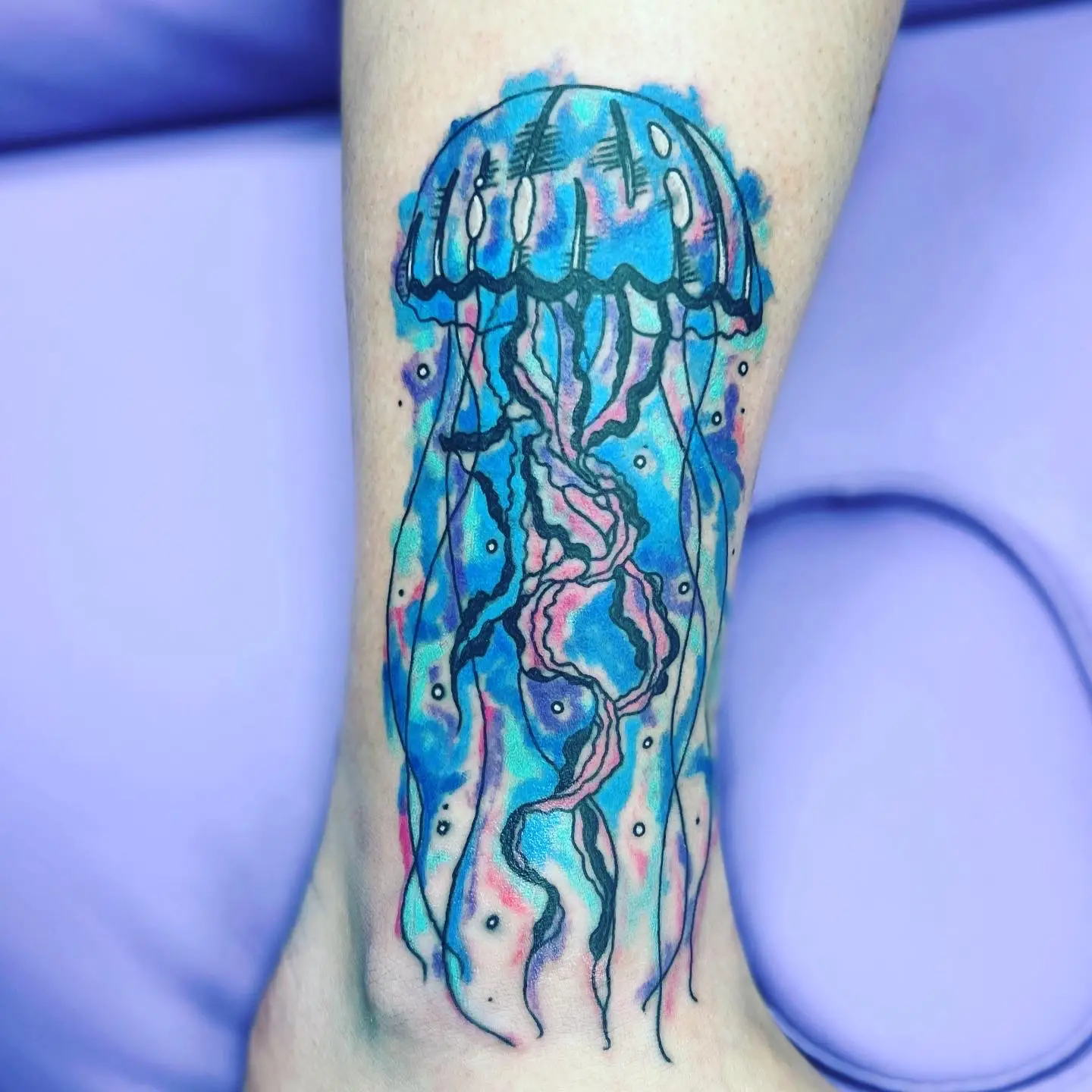 Watercolor jellyfish tattoo in bright blue, lavender, and pink with flowing tentacles on the lower leg