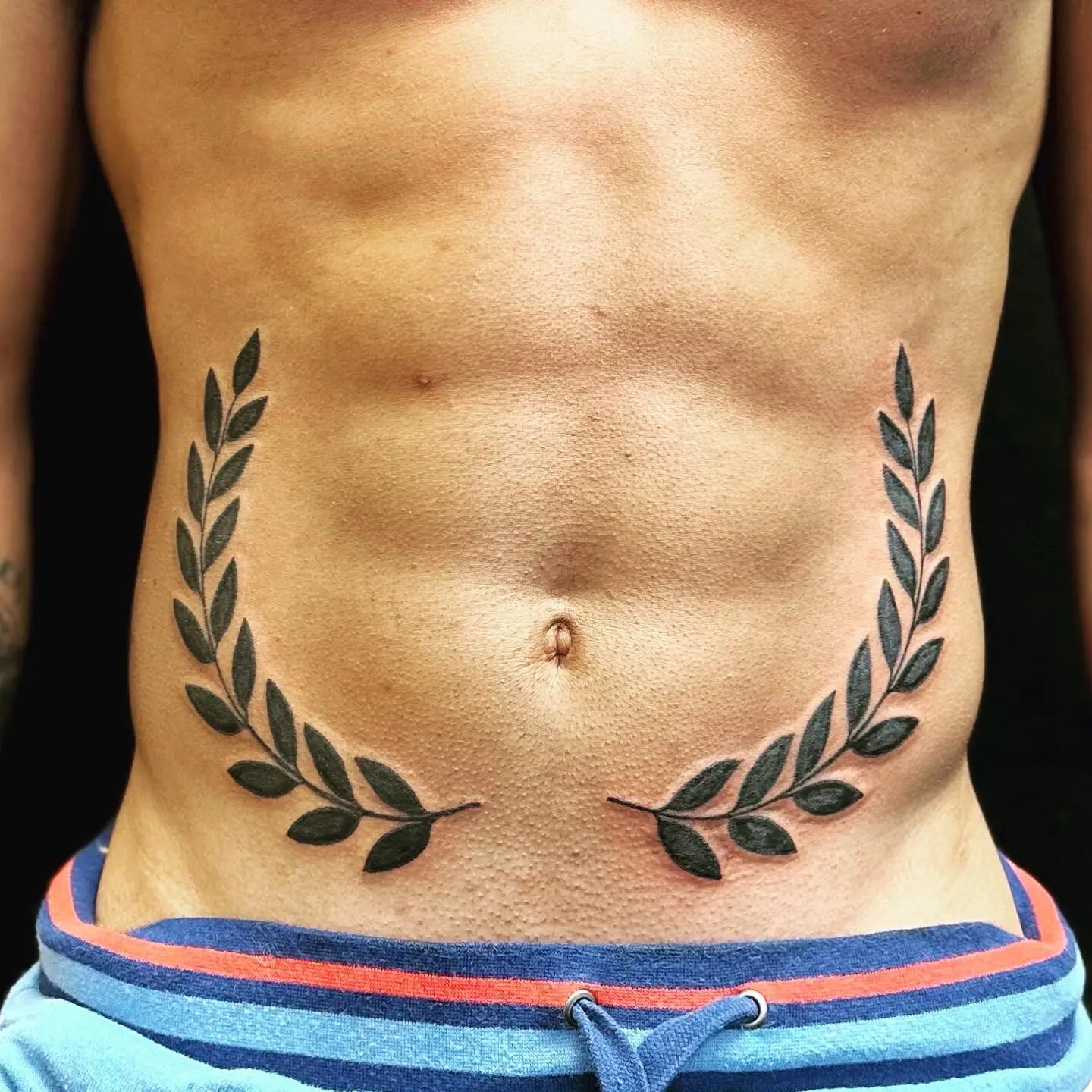 Bold black laurel wreath framing the navel across the stomach, illustrative tattoo by Carly