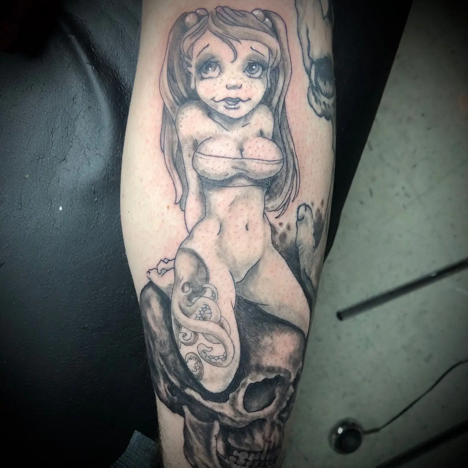 Black and grey calf tattoo of a wide-eyed cartoon mermaid resting on a skull entwined by an octopus