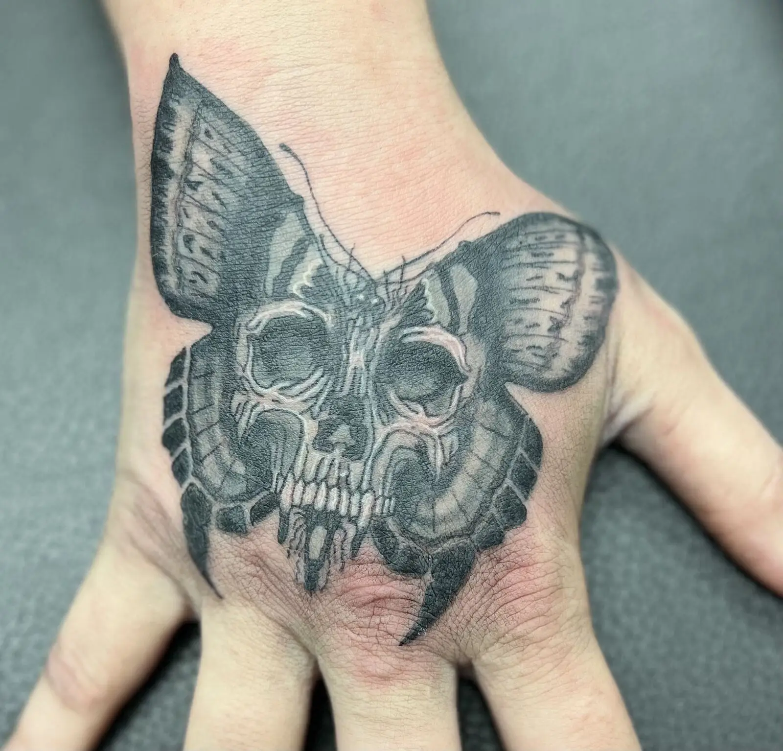 Black and grey hand tattoo combining a moth silhouette with a skull face and soft shading