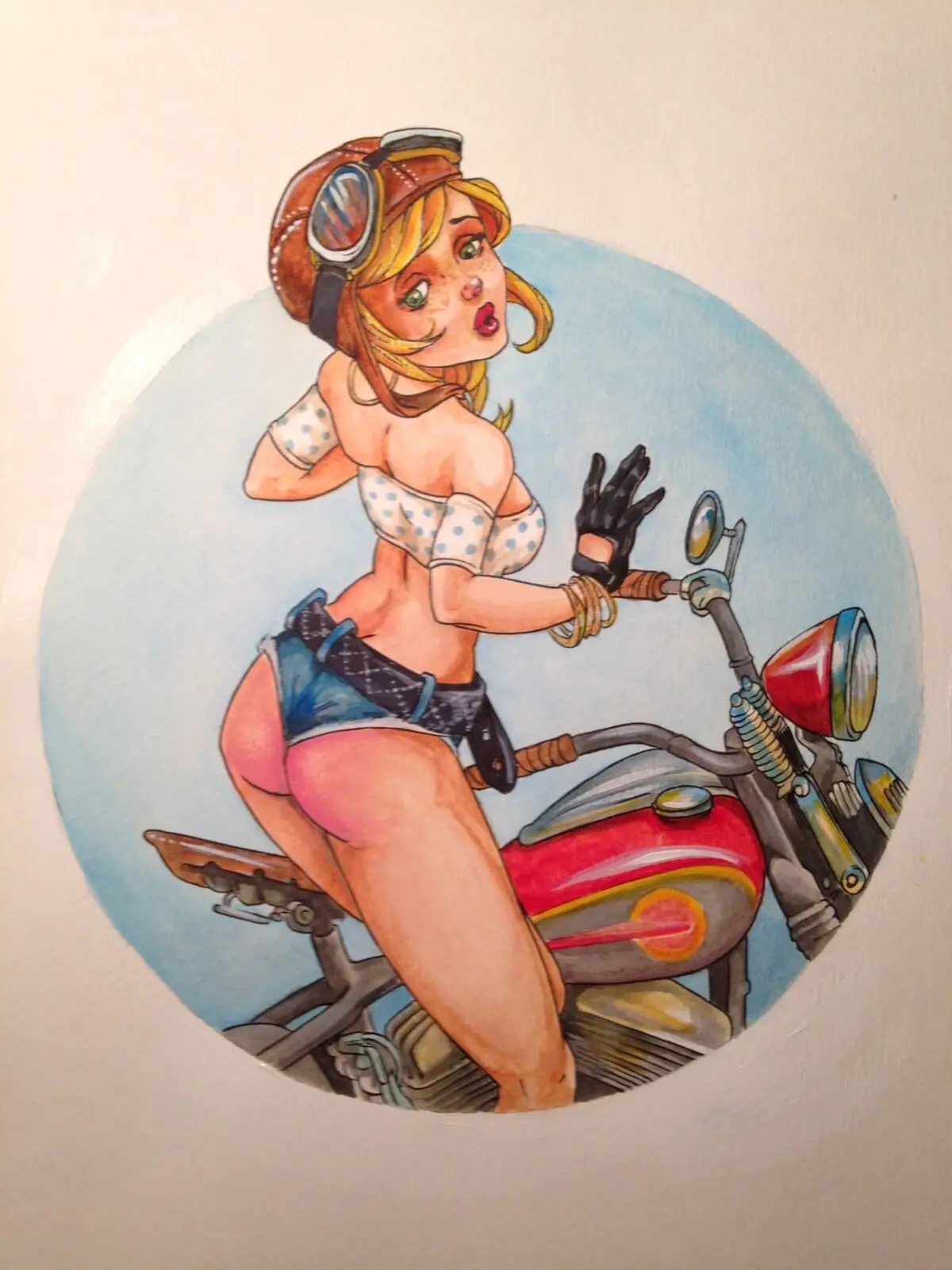 Watercolor pinup illustration of a freckled blonde biker girl in goggles and polka-dot top straddling a red vintage motorcycle inside a blue circle