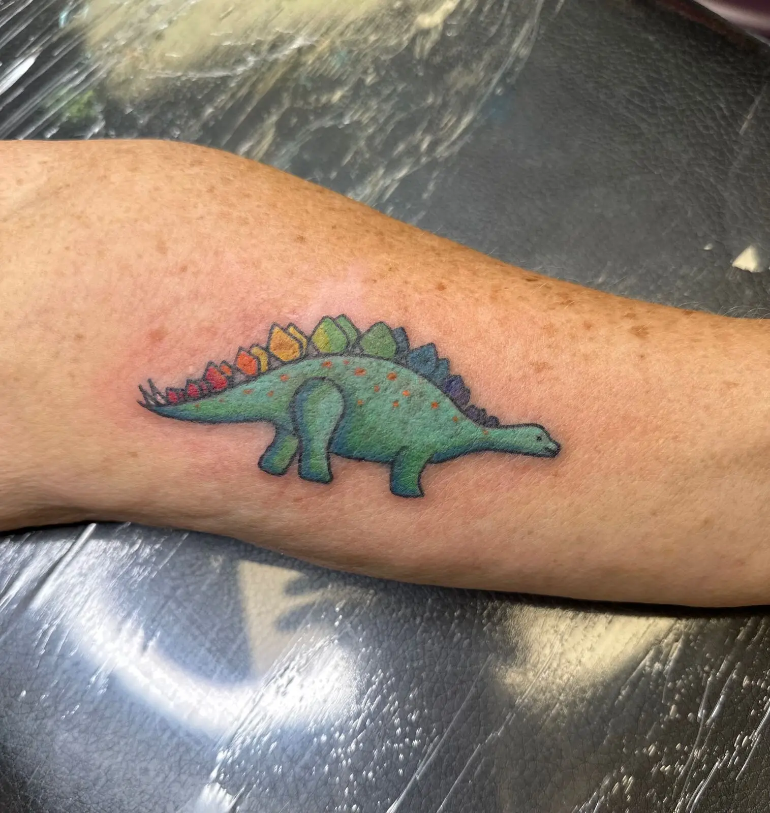 Playful color tattoo of a cartoon stegosaurus with rainbow plates and orange spots on the inner arm, by Carly