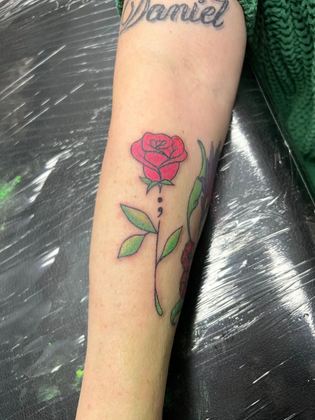 Color forearm tattoo of a red rose with a semicolon stem and green leaves, mental health awareness piece, by Carly