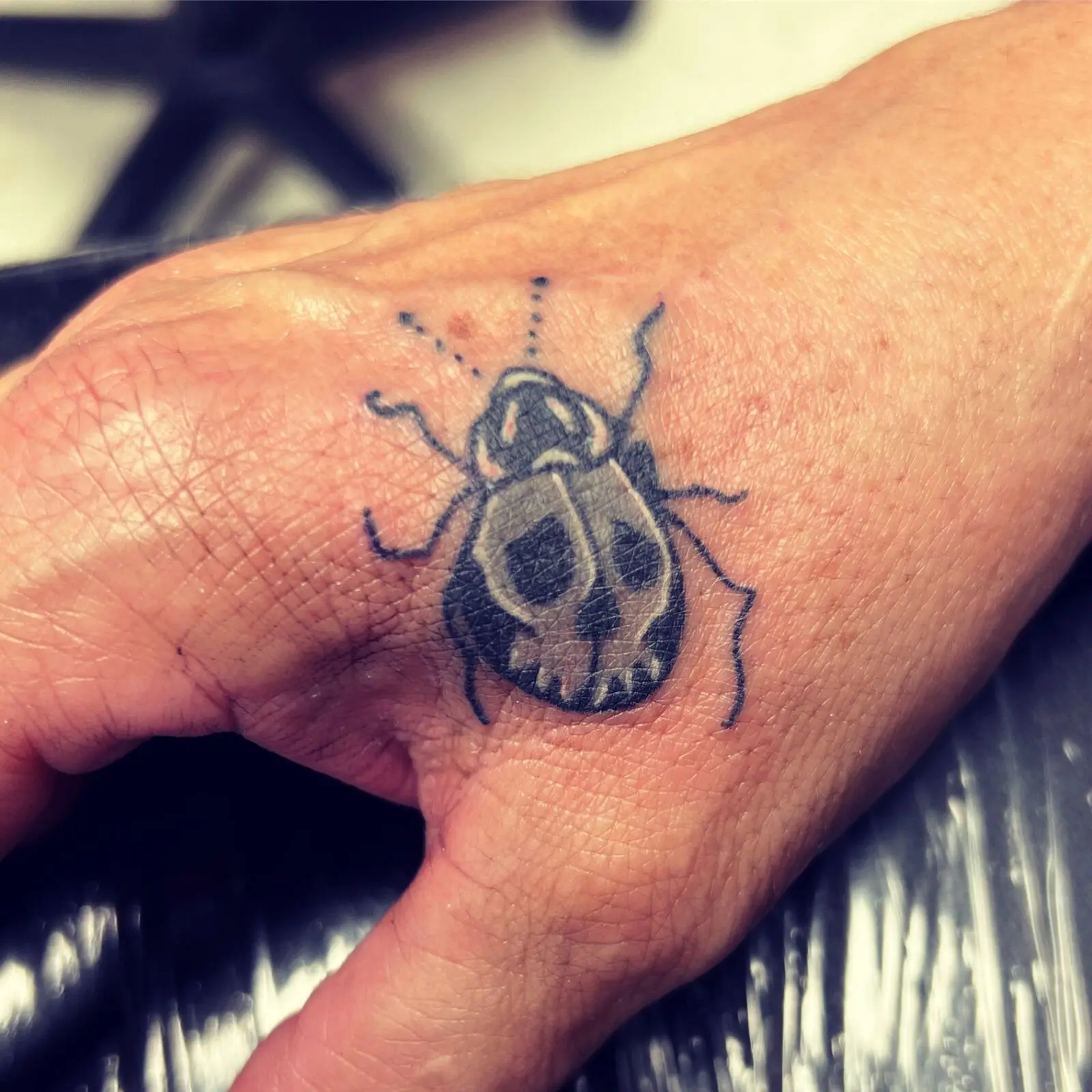 Small black and grey hand tattoo of a beetle with a skull-shaped shell and dotted accent marks