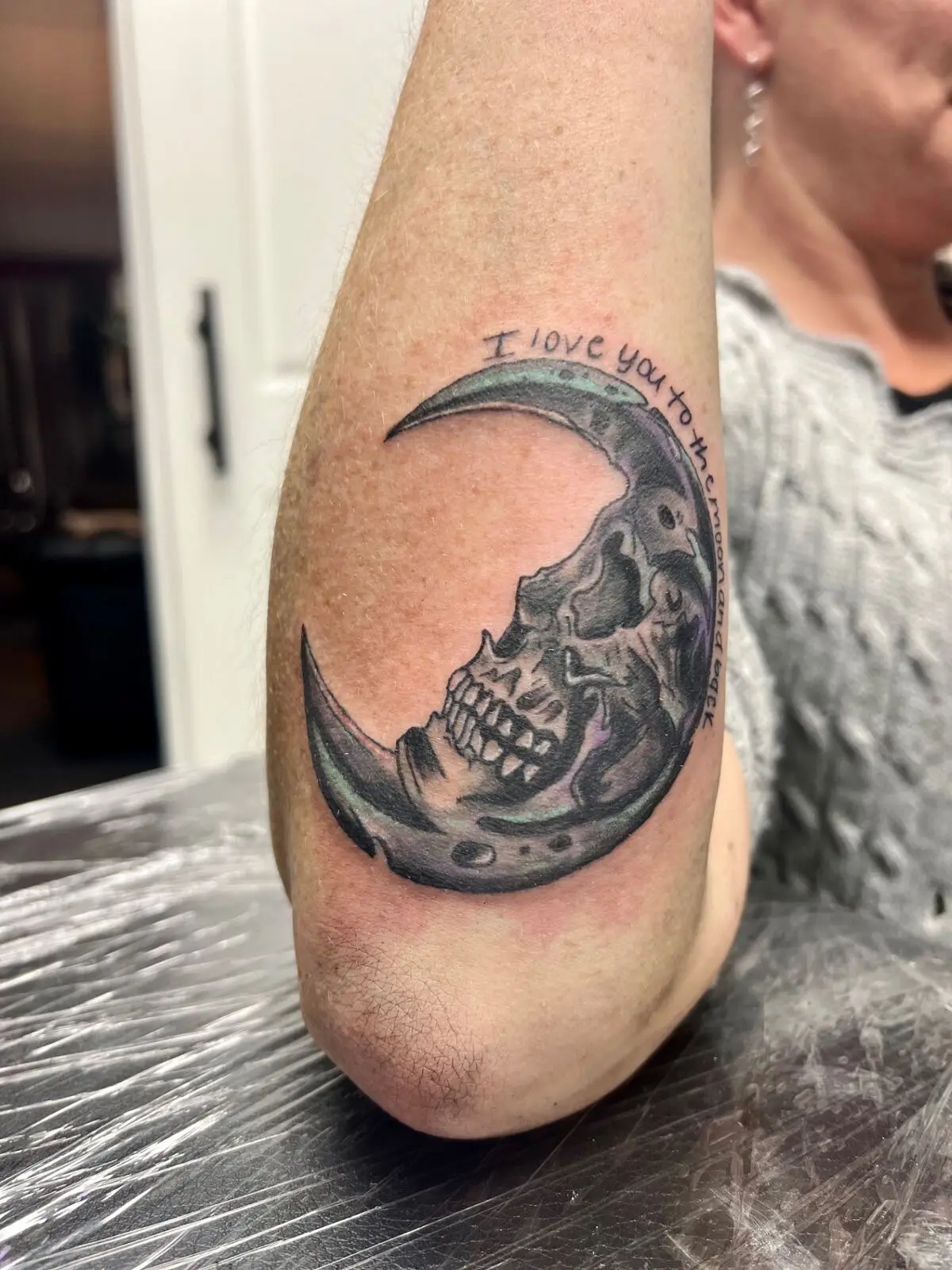 Color tattoo of a skull cradled in a teal and purple crescent moon under handwritten script reading I love you to the moon and back, by Carly