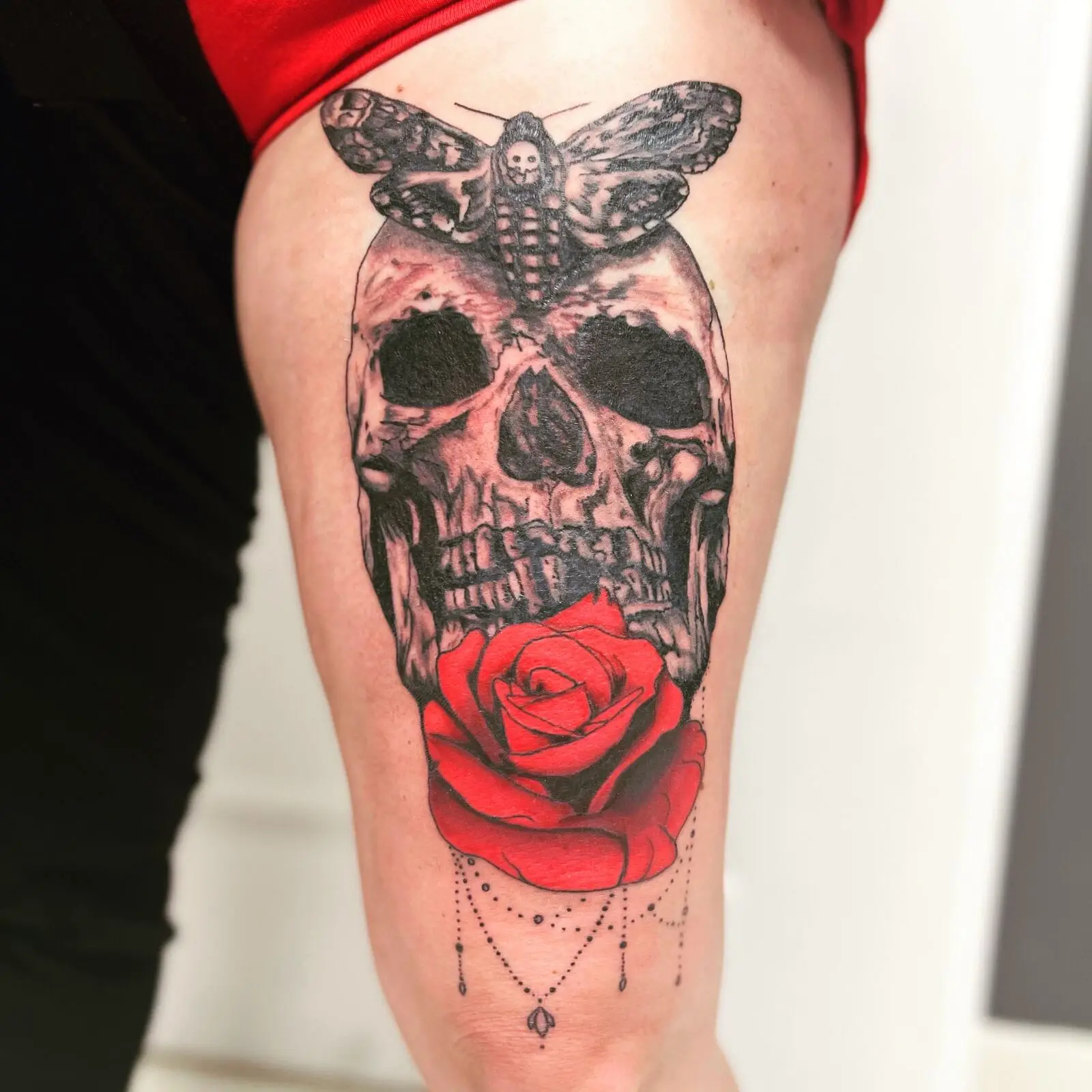 Large thigh tattoo of a realistic black and grey skull with a death's-head moth above and a vivid red rose with hanging beadwork below, by Carly