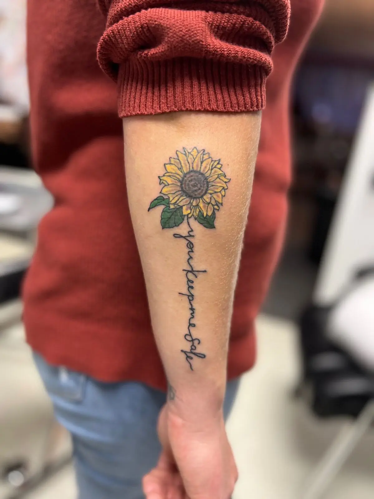 Fine line forearm tattoo featuring a sunflower above vertical script lettering reading You kept me safe