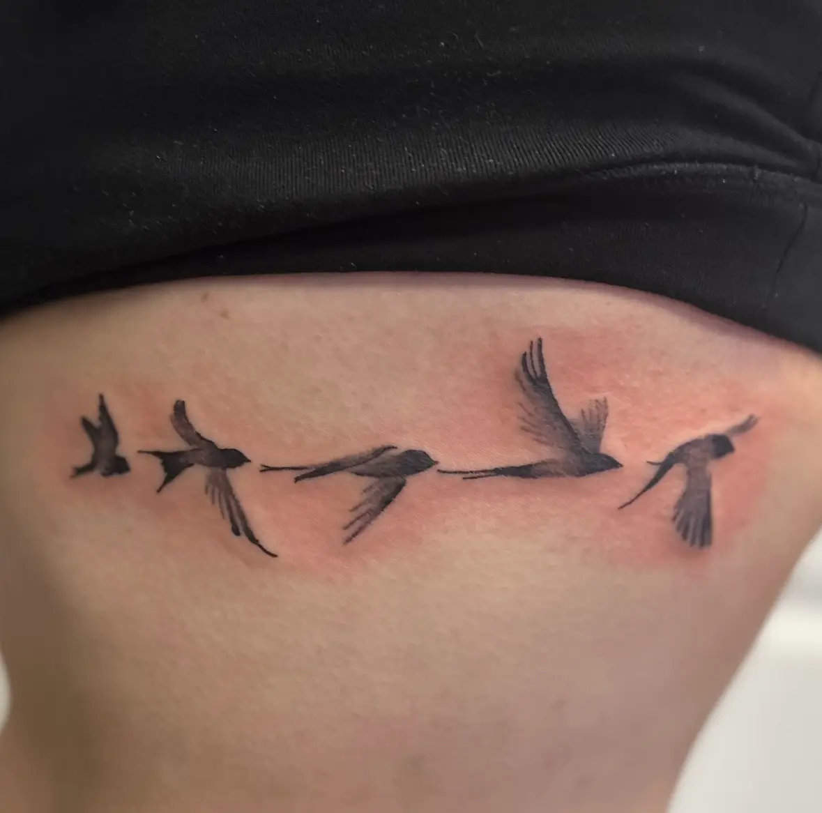 Delicate black and grey flock of swallows in flight across the ribs, fine line illustrative tattoo by Carly