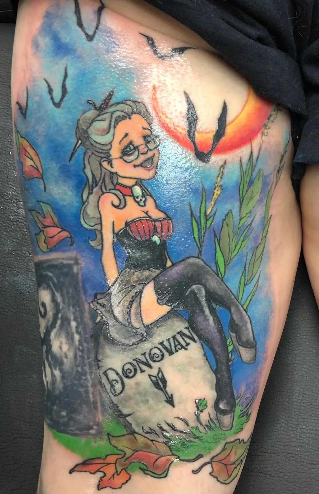 Color thigh tattoo of a cartoon pinup witch perched on a Donovan tombstone with bats and autumn leaves