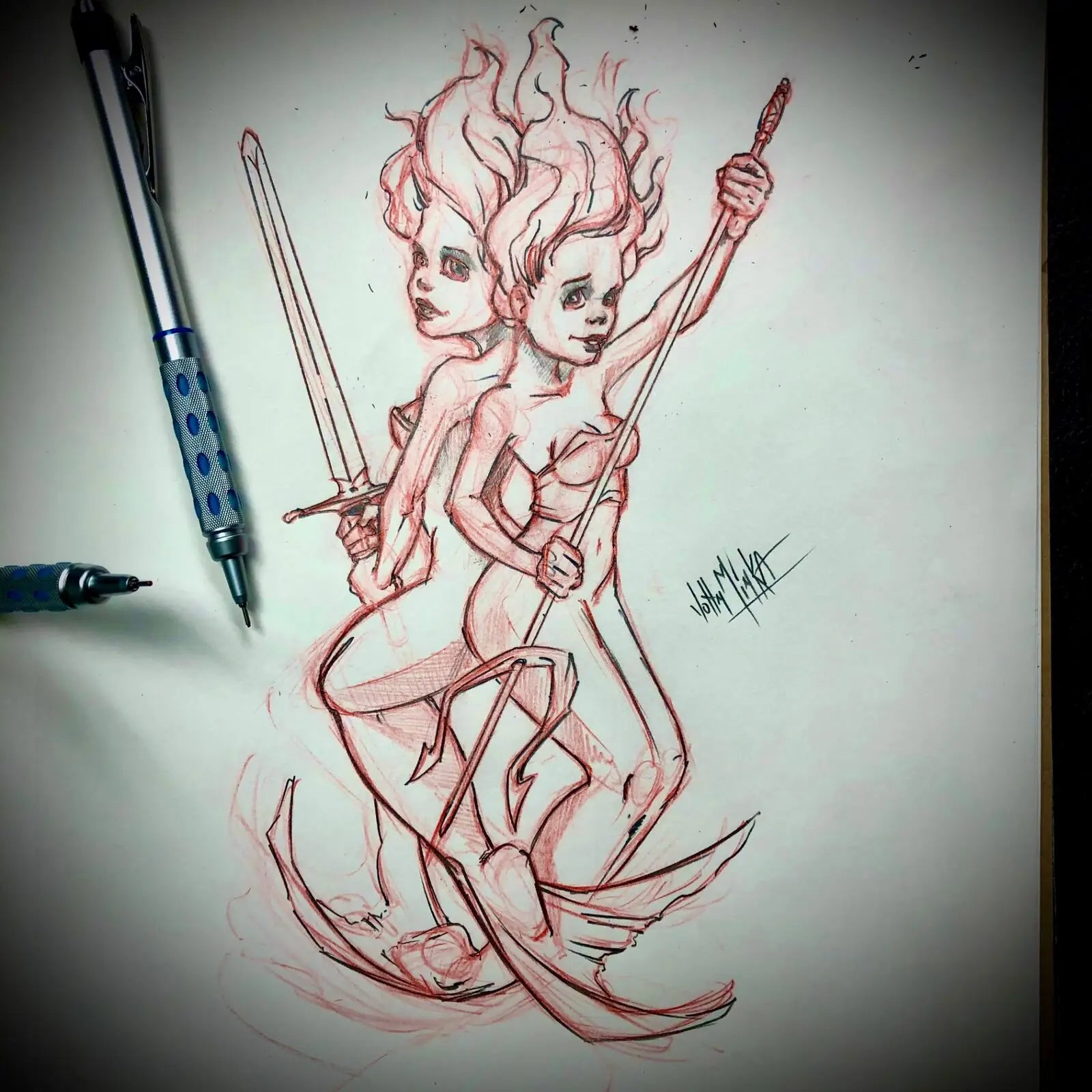 Red pencil sketch of a two-headed flame-haired warrior woman wielding a sword and bow, photographed beside drafting pens