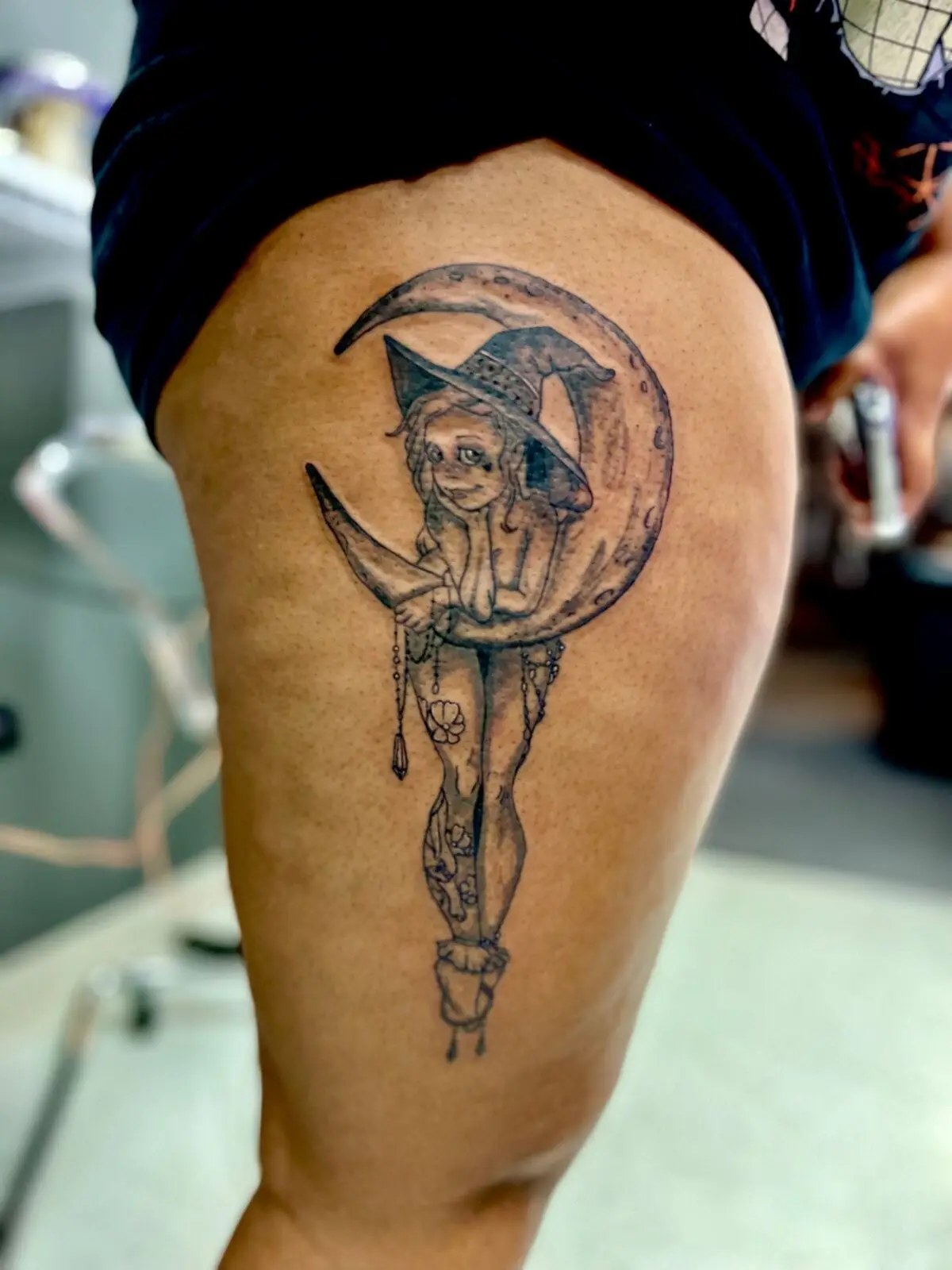 Black and grey illustrative thigh tattoo of a witch pinup in a wide brim hat lounging in a crescent moon with hanging crystals, by Carly